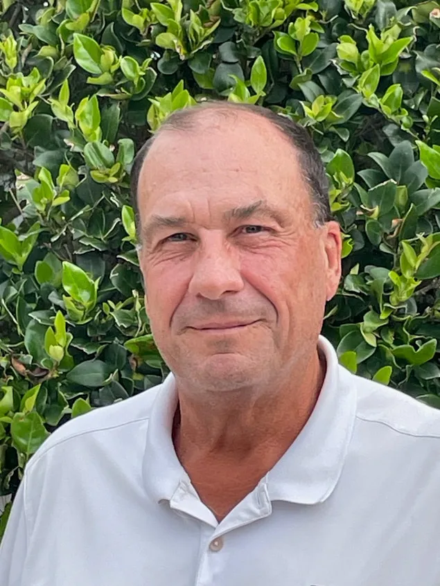 W. Kenneth Tyler, Jr., smiling in a white collared polo shirt in front of a green hedge.